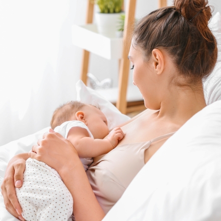 A mother in a beige camisole is lying in bed, gently cradling her baby while breastfeeding in a bright and cozy home setting.