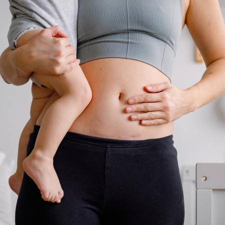 A mother in a gray sports bra and black leggings holds her baby while gently touching her postpartum abdomen in a softly lit home setting.